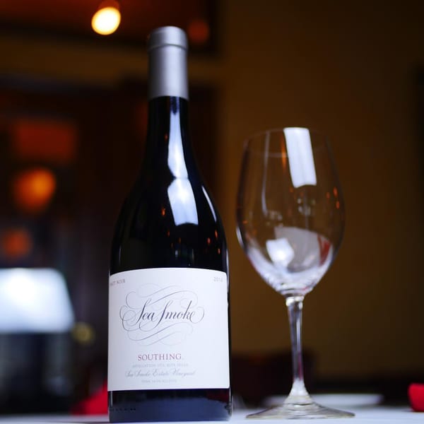 A bottle of Sea Smoke Pinot Noir wine stands next to an empty wine glass on a white tablecloth.