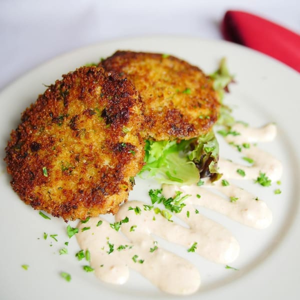 Two golden-brown Italian crab cakes with greens, creamy sauce, and parsley on a white plate.